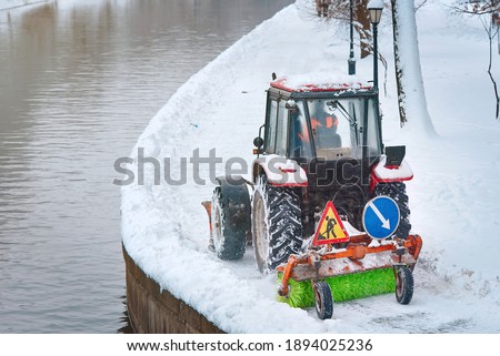 Tractor with snow plow and rotating brush sweeping snow from sidewalk on embankment in park. Red tractor with scoop and automated brush removes snow. Municipal road sweeping vehicle with plow Photo stock © 