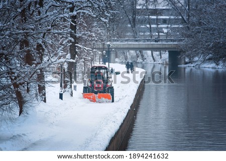 Tractor with snow plow and rotating brush sweeping snow from footpath on embankment in park. Red tractor with scoop and automated brush removes snow. Municipal road sweeping vehicle with plow Photo stock © 