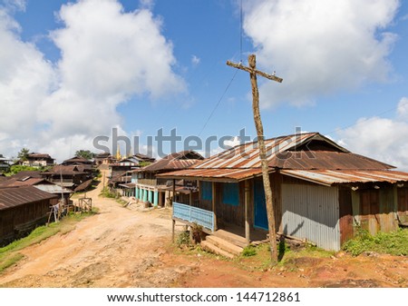 Mud bare earth village lane with wooden huts with corrugated sheet iron metal roofs and wooden makeshift electricity pole. A golden temple stupa at the end.