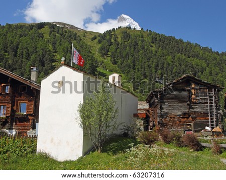 small swiss village settlement of wooden houses with little white stone church covered with stone roofes in front of forest and hugh snow covered alpine mountains on a sunny day in Wallis Switzerland