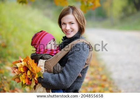 Young mother carrying her adorable baby daughter in a baby carrier on beautiful autumn park