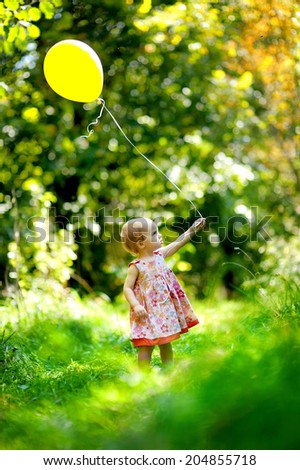 Adorable toddler girl holding yellow balloon having a walk in beautiful summer park