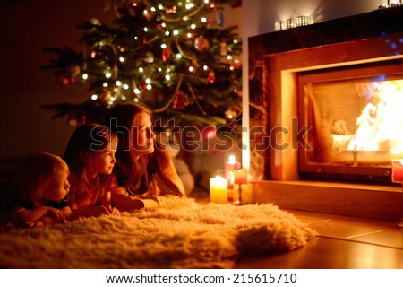 Young mother and her two little daughters sitting by a fireplace in a cozy dark living room on Christmas eve