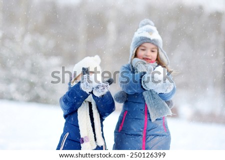 Two funny adorable little sisters having fun together in beautiful winter park during snowfall