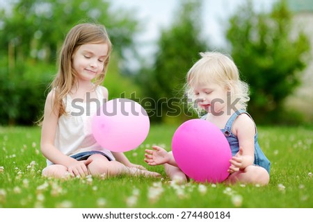 Two cute little sisters having fun with pink balloons while sitting on the grass on a sunny summer day