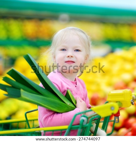 Cute little girl sitting in a shopping cart holding a leek in a food store