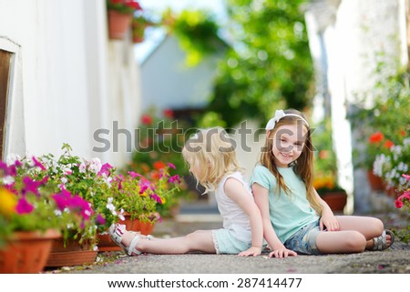 Two adorable little sisters sitting among flowers pots on warm and sunny summer day in italian town