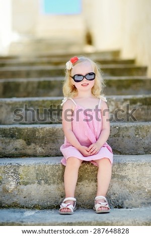 Adorable little girl wearing sunglasses sitting on stairs on warm and sunny summer day in typical italian town