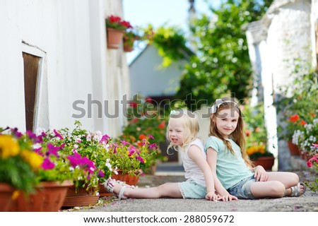 Two adorable little sisters sitting among flowers pots on warm and sunny summer day in italian town