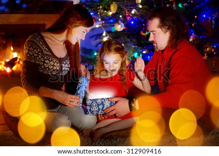 Young parents and their little daughter opening a magical Christmas gift by a Christmas tree in cozy living room in winter