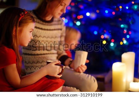 Young mother and her two little daughters drinking hot chocolate by a fireplace in a cozy dark living room on Christmas eve