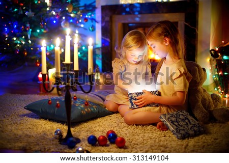 Adorable little girls opening a magical Christmas gift by a Christmas tree in cozy living room in winter