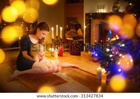 Young mother and her little daughter sitting by a fireplace in a cozy dark living room on Christmas eve