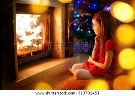 Happy little girl sitting by a fireplace in a cozy dark living room on Christmas eve