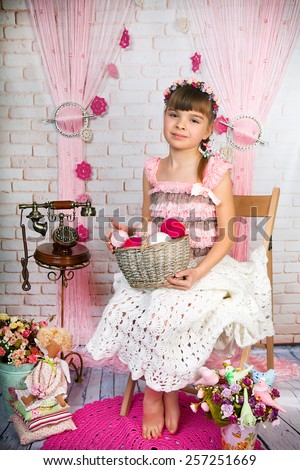 Cute little girl in pink fishnet dress and wreath holding a basket with balls of knitting sitting on a chair in the scenery