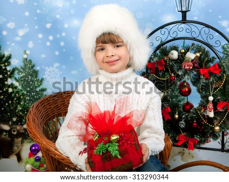 Portrait of a beautiful girl in a white cap and knit sweater sitting in wicker sleigh holding a gift on a background of Christmas decorations
