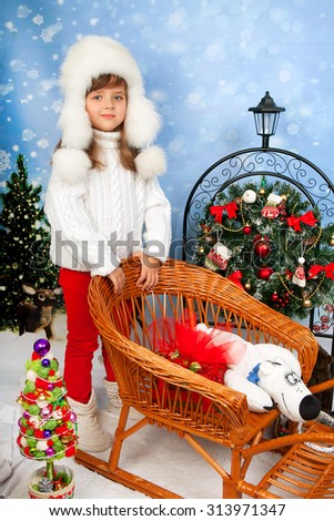 beautiful girl in a white cap and knit sweater costs about wicker sleigh with toys and gifts on the background of Christmas decorations