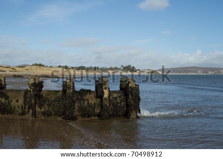Water drainage pipe made of metal with wooden supports on a beach, water is flowing into the sea, a popular beach destination in the distance.
