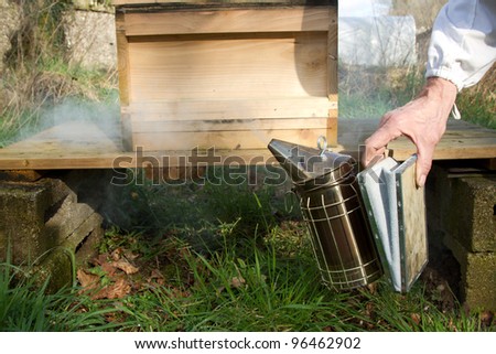 A metal bee smoker with bellows being squeezed by a hand in front of a wooden hive on a stand.