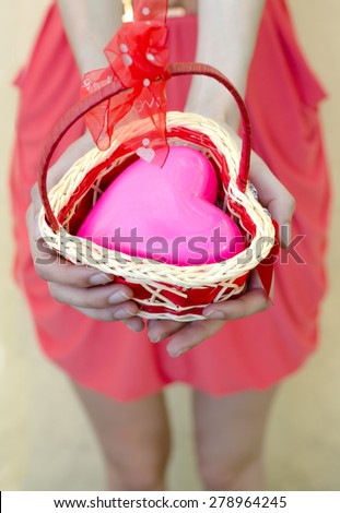 Female hands offering big pink heart in small knitted basket with ribbon.