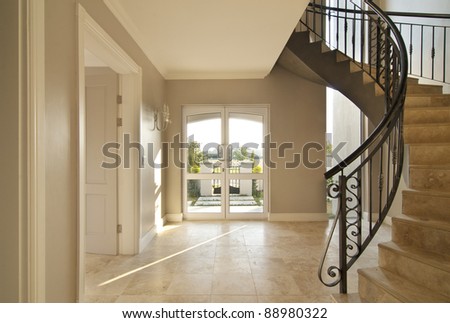 Staircase and front door area of a modern house. Staircase is framed on the right and the outside is visible through the front door. Both the staircase and floor is tiled