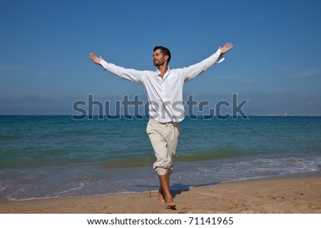 Happy handsome man smiling in front of the ocean with his arms open, wearing comfortable light clothes.