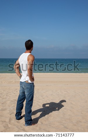 Muscular dark haired man looking at the ocean with his hands on his hips, wearing jeans and white shirt, viewed from behind.