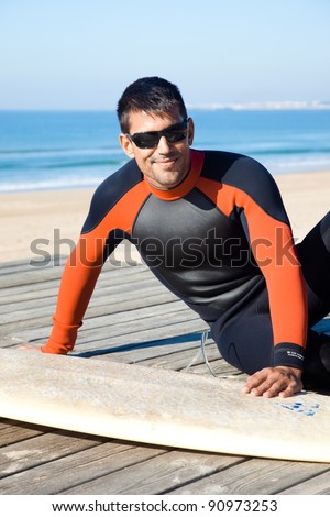 Handsome surfer wearing a wetsuit sitting next to his surf board smiling at camera.
