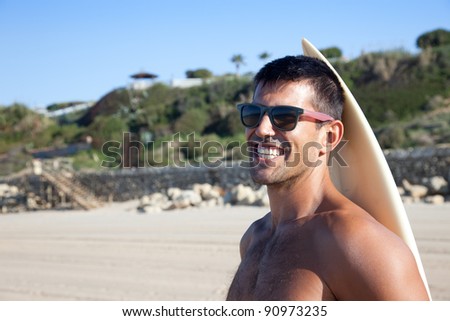 Surfer with sunglasses smiling at camera with his surf board behind him.