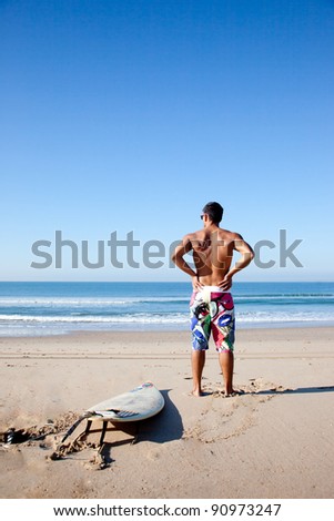 Surfer looking at the ocean (seen from behind) with his surf board on the sand next to him.