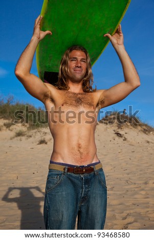 Fit long haired blonde surfer wearing jeans holding his green surfboard over his head.
