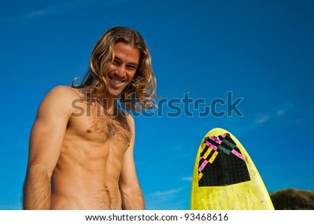 Long haired blonde surfer laughing next to his surfboard.