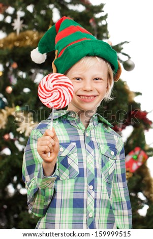 A cute boy dressed as Santa\'s helper or an elf is holding a red lollipop and smiling in front of a Christmas tree. Isolated on white.