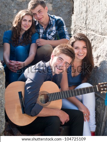 Group of four young happy smiling people with guitar having fun by posing outdoors in spring or summer sitting on the stone staircase.