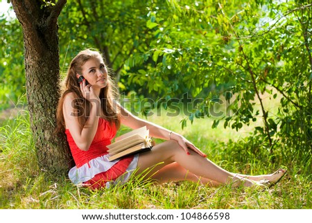 Portrait of a beautiful young woman in red sitting outdoors barefoot talking on the mobile phone and holding a book in the park or garden on a bright sunny day of summer on the green grass background