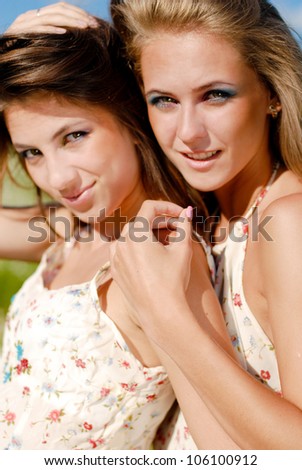 Two beautiful happy smiling young women embracing on blue sky and green grass background on a bright day of summer