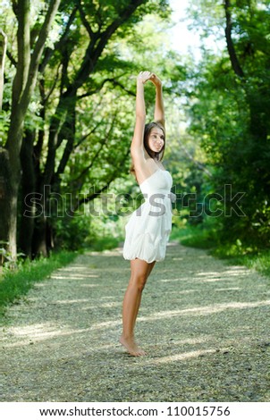 Young beautiful woman stretching hands up in white dress standing outdoors on the bright summer day in green park background