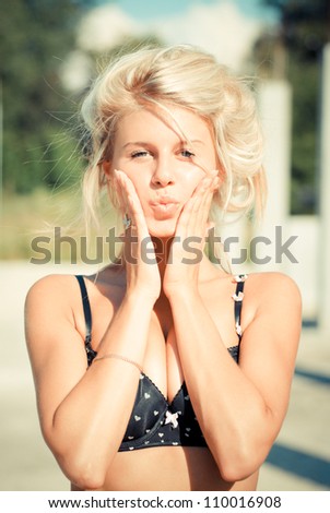 Young beautiful blond woman wearing bra touching cheeks and putting lips in kiss outdoors on the bright day of summer background