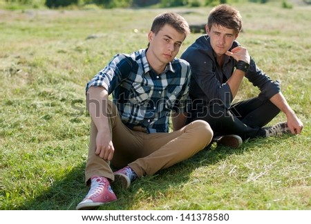 Two young men sitting together on green lawn on spring or summer day & green outdoors background