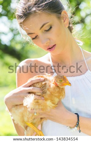 Happy young beautiful woman taking care of red chicken domestic bird on summer day
