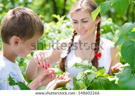 Teenage happy smiling sister and little brother eating raspberry outdoors on the summer day green outdoors background