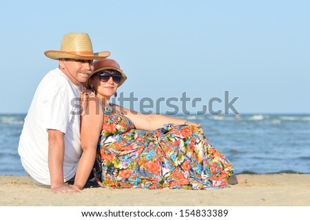happy smiling & looking at camera mature couple sitting at seashore on sandy beach back to back on the summer outdoors background
