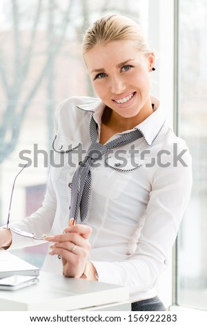 Beautiful business woman happy smiling & looking at camera holding glasses in office by window and computer
