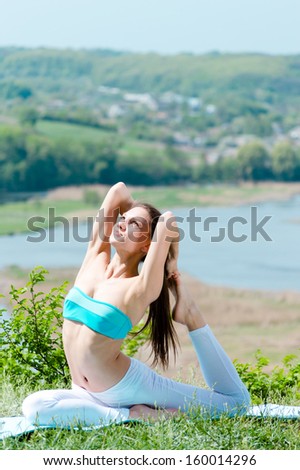 Happy young woman doing yoga on green river bank in summer outdoors