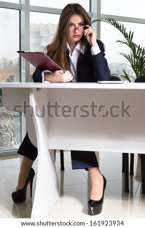 Successful business woman in man suit sitting at office table and looking over glasses