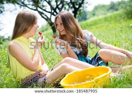 2 young happy smiling pretty women two teen girl friends having fun eating strawberry from huge bowl on summer day outdoors background