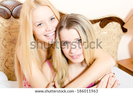 Two attractive blond young women beautiful girlfriends in pajamas having fun hugging sitting on white bed happy smiling & looking at camera closeup portrait