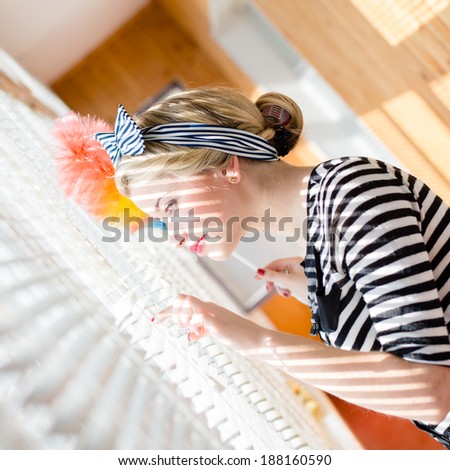 Blond pretty young woman holding a domestic dust stick looking at sunny window opening shutters