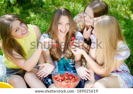 Four happy smiling beautiful group of young women teenagers having good time & fun eating strawberry on summer green outdoors background