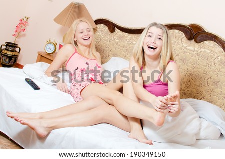 joyful foot massage treatment: beautiful blond girl friends having fun relaxing while younger sister making foot massage older sister on a white bed happy laughing looking at camera